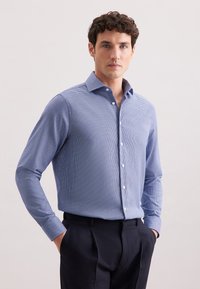 Uomo che indossa una camicia button-down azzurro chiaro a fantasia, slim-fit, infilata in pantaloni scuri su misura, con le mani in tasca, su uno sfondo neutro.