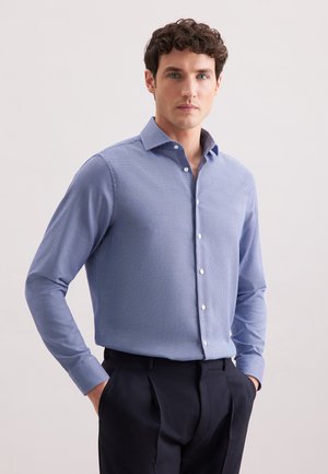 Uomo che indossa una camicia button-down azzurro chiaro a fantasia, slim-fit, infilata in pantaloni scuri su misura, con le mani in tasca, su uno sfondo neutro.