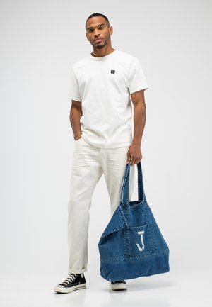 Mann trägt weißes T-Shirt und Hose, schwarze Sneakers, hält große blaue Denim-Tragetasche mit weißem "J" auf der vorderen Tasche.