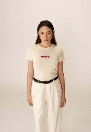 T-shirt couleur crème à manches courtes avec le mot "merci" en texte rouge, associé à un pantalon blanc taille haute et une ceinture noire.