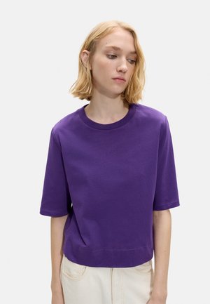 Młoda kobieta z blond włosami w boba, ubrana w prostą fioletową koszulkę z krótkim rękawem i jasne spodnie, patrząca w dół i lekko w lewo.