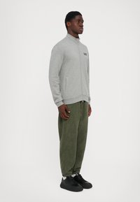Mann står med en grå zip-up jakke med EA7-logo, olivengrønne mønstrede joggebukser, hvite sokker og svarte sneakers på hvit bakgrunn.