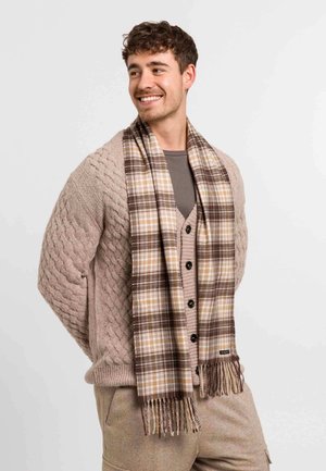 Uomo sorridente con cardigan beige a trecce, camicia marrone, pantaloni beige e sciarpa marrone a quadri su sfondo chiaro, mani dietro la schiena.