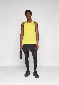 Gul atletisk tanktopp med teksturert mønster, svarte shorts, svarte leggings og sko. Holder en svart vannflaske.