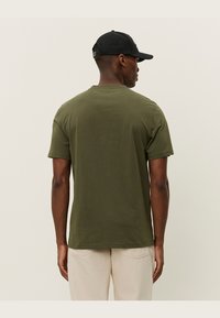 Olive green short-sleeve t-shirt, cotton material, round neck, relaxed fit, seamless shoulder design, worn with light-colored pants.