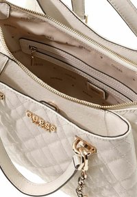 Sac à main blanc matelassé avec ferrures dorées et logo "GUESS". L'intérieur comprend une poche zippée et une doublure à motifs avec le nom de la marque.