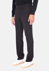 Uomo che indossa pantaloni neri su misura, una camicia nera a maniche lunghe e sneakers bianche con suola in gomma, in piedi di fronte a uno sfondo neutro.
