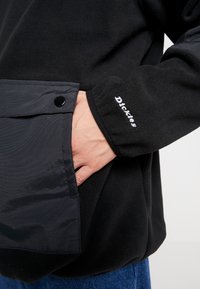 Chaqueta de forro polar negra con acabado texturizado, que cuenta con un bolsillo de nylon negro liso y un logo bordado en blanco. Corte ajustado con un bolsillo funcional.