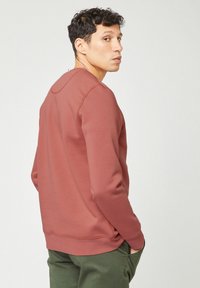 Uomo con capelli ricci e scuri che indossa una camicia a maniche lunghe color ruggine e pantaloni verdi, che guarda sopra la spalla con una mano in tasca.