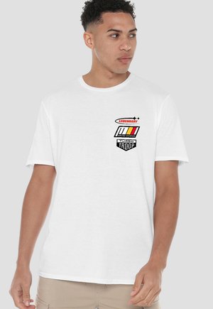 Young man wearing a white T-shirt with "Legendary," "Racing Division," and "Turbo Troop" logos in red, black, and yellow on the chest.