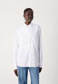White long-sleeve shirt with a button-down collar, chest pocket, and relaxed fit, made of smooth fabric. Paired with light blue jeans.