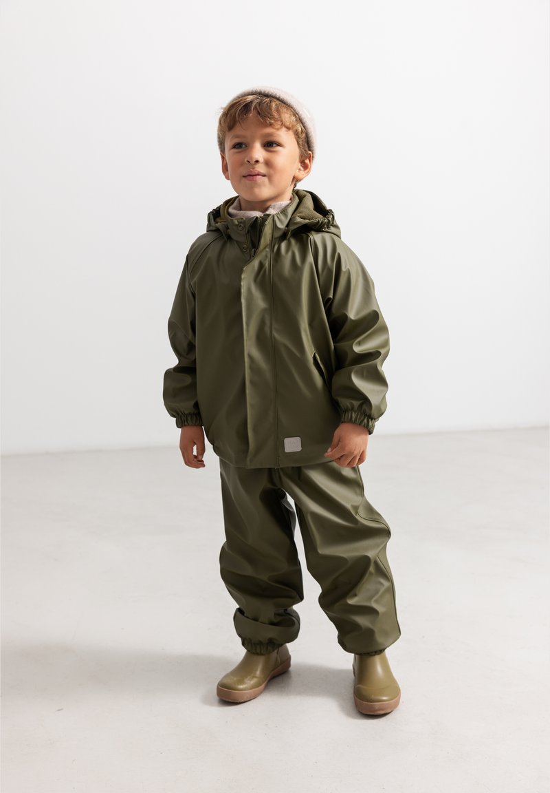 Jeune enfant portant une combinaison de pluie vert olive et des bottes, debout sur un sol clair avec un mur en arrière-plan neutre.