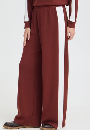 Personne portant un pantalon bordeaux à jambes larges avec une bande blanche sur le côté, haut assorti et chaussures brunes, debout sur un fond clair.