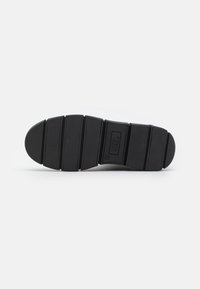 Suela de zapato de goma negra con un patrón estriado, que presenta seis ranuras horizontales y un logo central. Textura suave y diseño de punta redondeada.