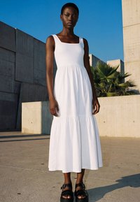 Eine große Frau in einem ärmellosen weißen Maxikleid und schwarzen Plateausandalen steht im Freien in der Nähe von Betonwänden und Palmenpflanzen unter klarem blauem Himmel.