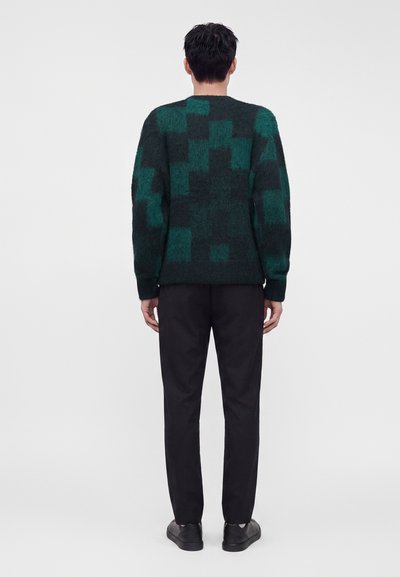 Homme de dos portant un pull patchwork vert et noir, un pantalon noir et des chaussures noires sur un fond clair uni.