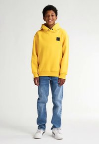 Sudadera amarilla con capucha, bolsillo frontal y logo, combinada con vaqueros azul claro y zapatillas blancas. Tejido de algodón, ajuste relajado, diseño sencillo.