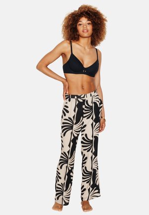 Vrouw staat op blote voeten met krullend haar en draagt een zwarte bh en beige wijde broek met een gedurfd zwart abstract bladerenprint.