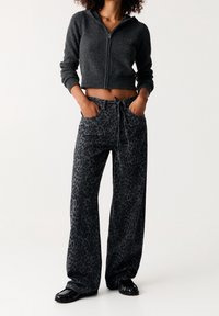 Sudadera gris de cremallera, recortada, con puños acanalados, combinada con pantalones negros de pierna ancha con un sutil estampado de leopardo. Unos zapatos negros completan el conjunto.