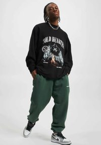 Melna sporta krekls ar grafisku apdruku, zaļas sporta bikses un melni un balti sporta apavi. Ievērojama brīva piegriezuma un izteikts kakla dizains.