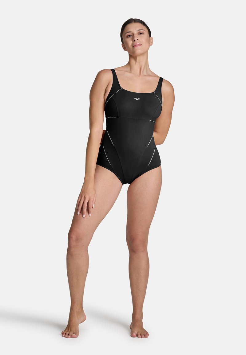 Femme debout pieds nus dans un maillot de bain noir une pièce avec des coutures blanches subtiles sur fond blanc.