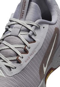 Nike Trail running shoe in light gray mesh with textured accents, featuring a white logo, brown details, and a durable sole with splatter pattern.