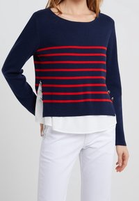 Navy sweater with red horizontal stripes, ribbed texture. Features side slit and decorative metal accents. Layered over white shirt.