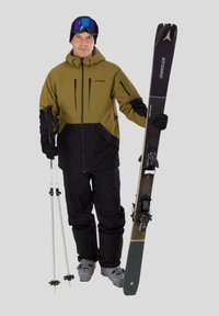 Lyžařský outfit: olivově zelená a černá bunda se zipy, černé kalhoty, šedé lyžařské boty, lyžařské hole a lyže s tmavým designem a vázáním.