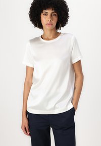 T-shirt z krótkim rękawem w kolorze białym, wykonany z gładkiego materiału, z okrągłym dekoltem i luźnym krojem, zestawiony z ciemnymi spodniami.