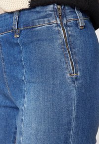 Jeans de mezclilla azul con una textura suave, que presentan un cierre lateral, diseño de cinco bolsillos y costuras amarillas en contraste a lo largo de las costuras.
