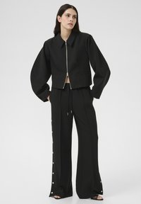 Zwarte cropped jas met rits en twee frontzakken, gecombineerd met zwarte broeken met wijde pijpen en decoratieve knopen langs de zijkanten.