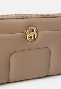 Detailný záber na béžovú koženú tašku so zlatým emblémom a viditeľným šitím, vybavenú zipsom na vrchu.
