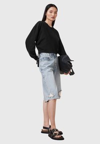Sudadera negra recortada, pantalones cortos de mezclilla desgastados de color azul claro y sandalias negras con acentos en dorado. Modelo sostiene un bolso negro.