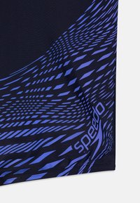 Ναυτικές μαγιό σορτς με γεωμετρικό μπλε μοτίβο. Το ύφασμα έχει λεία υφή. Το λογότυπο "Speedo" είναι ορατό στο πλάι.