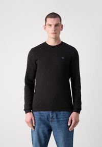 Svart langermet skjorte med et teksturert mønster, rund hals og en liten logo på brystet. Kombinert med blå denimjeans.