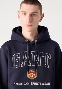 Sudadera azul marino de tejido suave con gran texto "GANT" y un logo de escudo que incluye "USA" y "1949" en colores contrastantes.