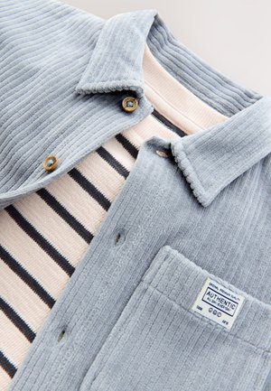 Chemise en velours côtelé bleu clair avec deux boutons en bois et une poche, superposée à un t-shirt à col rond rayé rose. Tissu texturé.