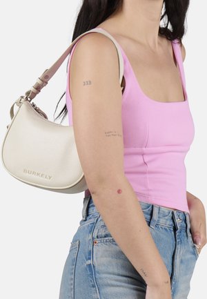 Crème gekleurde leren handtas met een gebogen vorm, gouden hardware en een verstelbare schouderband, gepresenteerd tegen een roze top en spijkerbroek.
