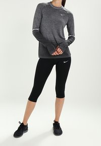 Dlouhý rukáv sportovního trička Nike v tmavě šedé barvě s texturovaným vzorem, spárovaný s černými legínami a černými sportovními botami.