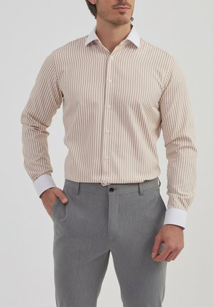 Chemise boutonnée à rayures marron clair et blanche, manches longues avec des poignets blancs contrastés, portée avec un pantalon gris, design ajusté.