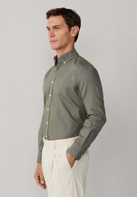 Mężczyzna w oliwkowozielonej koszuli zapinanej na guziki z kołnierzem i kremowych plisowanych spodniach, stojący z jedną ręką w kieszeni na jednolitym tle.