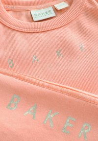 Peachfärgad, ribbad bomullströja med silverfärgad text "BAKER", rund halsringning och etiketter som säger "BAKER by TED BAKER."