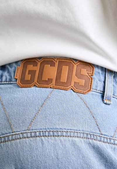 Denim kavbojke svetlo modre barve z vidnimi šivi, z rjavim usnjenim obližem s tiskanjem "GCDS" na pasu.