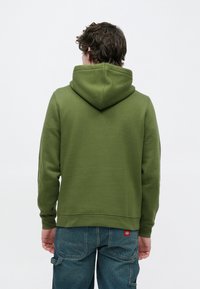 Πράσινο φούτερ με κουκούλα σε χαλαρή εφαρμογή, κατασκευασμένο από απαλό ύφασμα. Διαθέτει τσέπη καγκουρό και ριμπ μανσέτες. Φορεμένο με μπλε παντελόνια cargo.