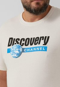 Beige Baumwoll-T-Shirt mit dem Aufdruck "Discovery Channel" in fetter schwarzer Schrift mit blauen Akzenten und einer blau-weißen Erdkugelgrafik.