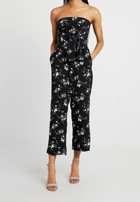 Mono floral negro sin mangas, con diseño de nudo en la cintura, bolsillos y pierna acortada, hecho de una tela ligera.