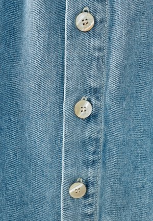 Svijetloplava traper tkanina s vertikalnim redom od četiri okrugla, biserna dugmeta, koja imaju teksturiranu površinu i šivane detalje.