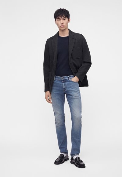 Homme portant un blazer noir, une chemise bleu marine foncé, un jean bleu clair et des mocassins en cuir noirs, debout avec une main dans la poche sur fond blanc.