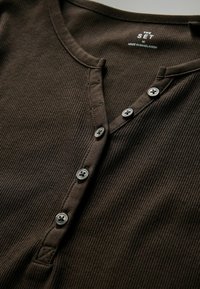 Pruun ribaga kootud särgi ümar kaelus, viie nupuga ja sildiga, millel on kiri "The Set". Kangas tundub pehme ja tekstuurne.