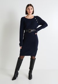 Vestido de punto azul marino con mangas largas, que presenta una textura de canalé y tres botones dorados en el hombro, ceñido a la cintura con un cinturón negro.